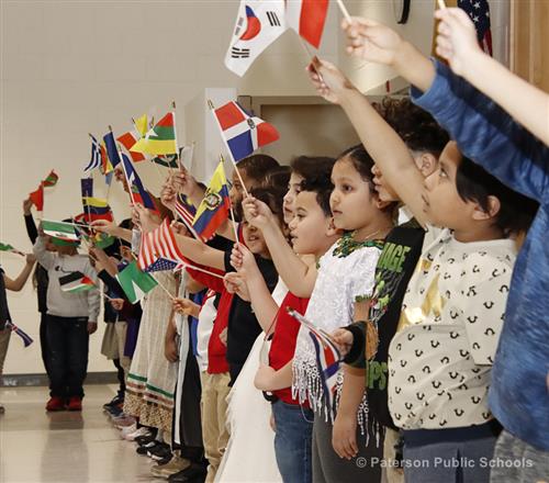 A long line of young students wave miniature flags. Students are lined up on the right-hand side towards center frame.